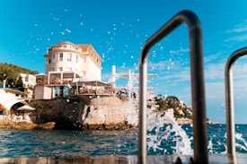 A picturesque view of a coastal area featuring a large, elegant villa perched atop a rocky cliff. The villa is surrounded by lush greenery and overlooks a sparkling body of water. Splashes of water are captured in motion against a clear blue sky. In the foreground, metal railings are visible, possibly indicating a seaside promenade or dock.