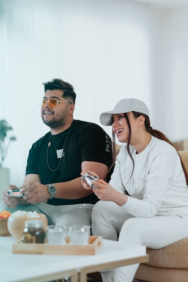 A man and a woman are sitting side by side on a couch, playing video games. The man is wearing a black shirt and glasses, while the woman is dressed in a white outfit and a cap. They both appear to be enjoying their time, smiling and engaged in the game. On the table in front of them, there are some snacks and drinks.