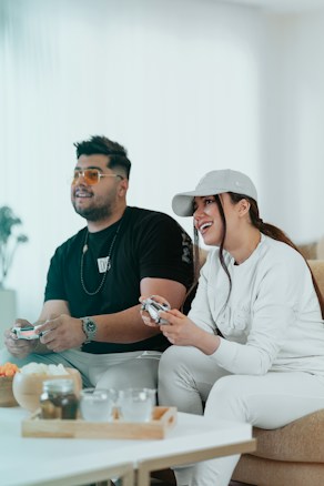 A man and a woman are sitting side by side on a couch, playing video games. The man is wearing a black shirt and glasses, while the woman is dressed in a white outfit and a cap. They both appear to be enjoying their time, smiling and engaged in the game. On the table in front of them, there are some snacks and drinks.
