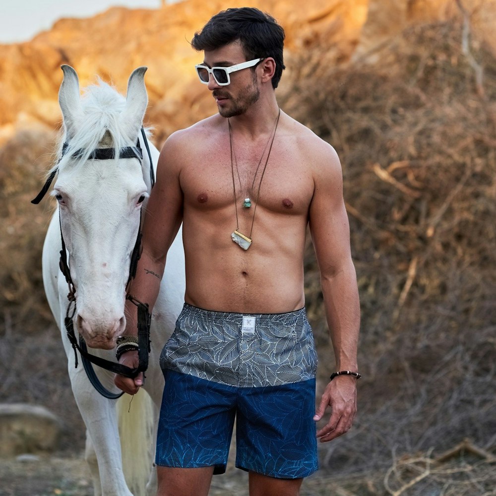Hombre en topless con pantalones cortos de mezclilla azul de pie junto a un caballo blanco durante el día