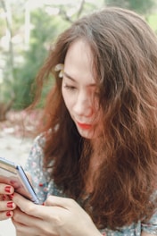 A woman with wavy brown hair is looking at a smartphone. She is wearing a floral-patterned shirt, and there is a white flower tucked behind her ear. Her nail polish is bright red, and the background is slightly blurred with greenery.