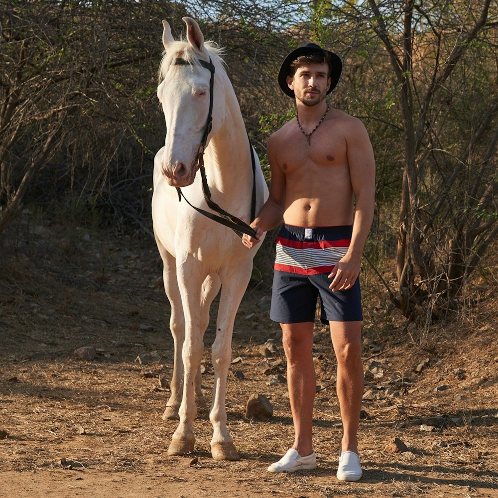 topless man in blue shorts standing beside white horse during daytime