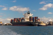 A large cargo ship is seen carrying numerous multicolored shipping containers stacked high on its deck. The water around the ship is calm, and a smaller yellow and black tugboat is assisting the cargo ship. The background shows an industrial port area with cranes and buildings under a partly cloudy sky.