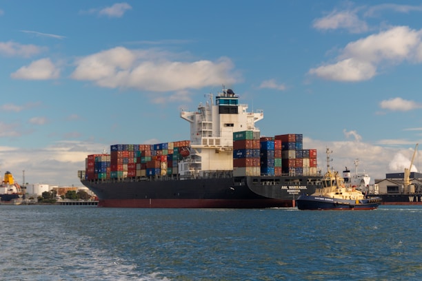 A large cargo ship is seen carrying numerous multicolored shipping containers stacked high on its deck. The water around the ship is calm, and a smaller yellow and black tugboat is assisting the cargo ship. The background shows an industrial port area with cranes and buildings under a partly cloudy sky.