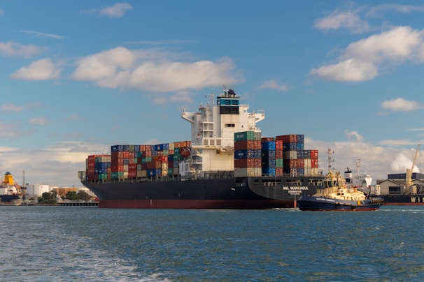 A large cargo ship is seen carrying numerous multicolored shipping containers stacked high on its deck. The water around the ship is calm, and a smaller yellow and black tugboat is assisting the cargo ship. The background shows an industrial port area with cranes and buildings under a partly cloudy sky.