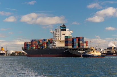 A large cargo ship is seen carrying numerous multicolored shipping containers stacked high on its deck. The water around the ship is calm, and a smaller yellow and black tugboat is assisting the cargo ship. The background shows an industrial port area with cranes and buildings under a partly cloudy sky.