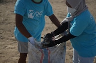Two individuals wearing matching blue t-shirts, face masks, and gloves are collaborating on collecting waste into a large woven sack on a sandy surface, suggesting an activity related to beach cleanup or environmental conservation.