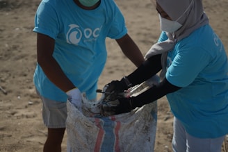 Two individuals wearing matching blue t-shirts, face masks, and gloves are collaborating on collecting waste into a large woven sack on a sandy surface, suggesting an activity related to beach cleanup or environmental conservation.
