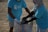 Two individuals wearing matching blue t-shirts, face masks, and gloves are collaborating on collecting waste into a large woven sack on a sandy surface, suggesting an activity related to beach cleanup or environmental conservation.