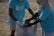 Two individuals wearing matching blue t-shirts, face masks, and gloves are collaborating on collecting waste into a large woven sack on a sandy surface, suggesting an activity related to beach cleanup or environmental conservation.
