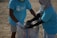 Two individuals wearing matching blue t-shirts, face masks, and gloves are collaborating on collecting waste into a large woven sack on a sandy surface, suggesting an activity related to beach cleanup or environmental conservation.