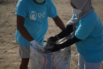 Two individuals wearing matching blue t-shirts, face masks, and gloves are collaborating on collecting waste into a large woven sack on a sandy surface, suggesting an activity related to beach cleanup or environmental conservation.