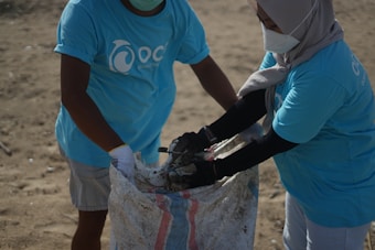 Two individuals wearing matching blue t-shirts, face masks, and gloves are collaborating on collecting waste into a large woven sack on a sandy surface, suggesting an activity related to beach cleanup or environmental conservation.