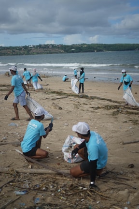 A group of people wearing matching blue shirts and caps are cleaning a beach, collecting debris and trash into large bags. The scene takes place on a sandy beach with the ocean in the background, surrounded by a cloudy sky.