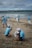 A group of people wearing matching blue shirts and caps are cleaning a beach, collecting debris and trash into large bags. The scene takes place on a sandy beach with the ocean in the background, surrounded by a cloudy sky.