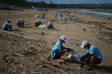 Several individuals wearing blue shirts and white caps are participating in a beach cleanup. They are collecting rubbish and debris scattered across the sand. The background includes the ocean with gentle waves and distant greenery or hills.