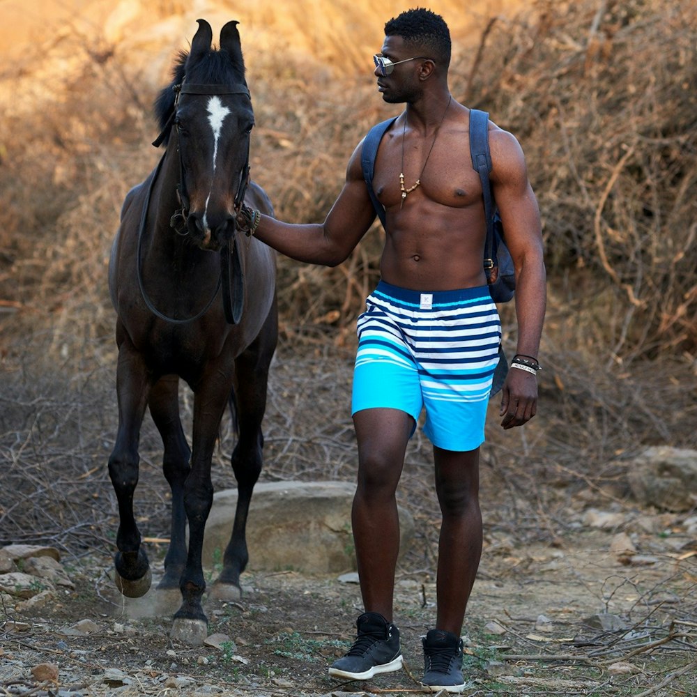 topless man in blue and white shorts standing beside brown horse during daytime