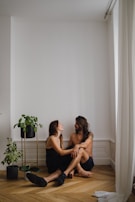 A couple sits on the wooden floor of a minimalistic room with white walls and curtains. They are smiling at each other, displaying a sense of intimacy and connection. Two potted plants are placed beside them, adding a touch of greenery to the serene environment.