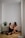 A couple sits on the wooden floor of a minimalistic room with white walls and curtains. They are smiling at each other, displaying a sense of intimacy and connection. Two potted plants are placed beside them, adding a touch of greenery to the serene environment.