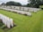 Rows of neatly aligned white headstones spread across a lush green cemetery. Vibrant red flowers and small shrubs are planted beside some of the headstones. The scene is peaceful and solemn, bordered by mature trees and a hedgerow, under a cloudy sky.