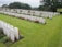 Rows of neatly aligned white headstones spread across a lush green cemetery. Vibrant red flowers and small shrubs are planted beside some of the headstones. The scene is peaceful and solemn, bordered by mature trees and a hedgerow, under a cloudy sky.