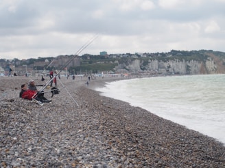 A pebble beach with a couple of people seated and fishing with long fishing rods. The beach extends into the distance with more people walking and scattered fishing poles. The sea waves are gently hitting the shore, and there are white cliffs with scattered buildings in the background. The sky is mostly cloudy with a few patches of blue.