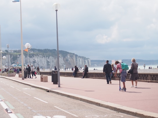 People walking and engaging in leisure activities along a seaside promenade with white cliffs in the background. The sky is overcast, and there are people on a beach and using scooters. Street lamps and barriers line the walkway.