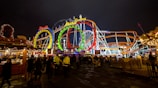 Brightly lit amusement park with colorful roller coaster loops and a lively crowd beneath the night sky. The scene includes vibrant signs and decorative stalls, creating a festive atmosphere.