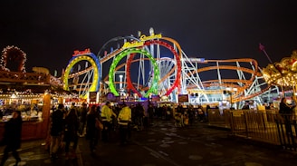 Brightly lit amusement park with colorful roller coaster loops and a lively crowd beneath the night sky. The scene includes vibrant signs and decorative stalls, creating a festive atmosphere.