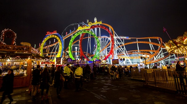 Brightly lit amusement park with colorful roller coaster loops and a lively crowd beneath the night sky. The scene includes vibrant signs and decorative stalls, creating a festive atmosphere.