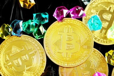 Several shiny gold coins featuring the Bitcoin symbol surrounded by brightly colored gemstones.