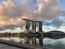 A modern cityscape featuring an iconic hotel with three towers connected by a unique rooftop structure, set against a dramatic sky with large, fluffy clouds. The foreground shows a calm body of water reflecting the architecture, with subtle lighting suggesting an early morning or late afternoon scene.
