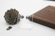 A pair of skull-themed earrings displayed on a transparent stand is set against a backdrop of a large pinecone and a dark wooden surface. The scene appears minimalist with a clean, white background, emphasizing the earthy tones of the wood and the textured pinecone.