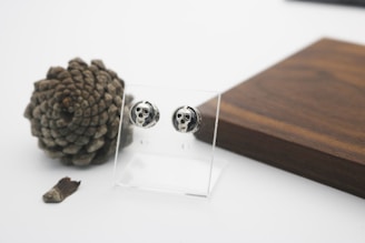 A pair of skull-themed earrings displayed on a transparent stand is set against a backdrop of a large pinecone and a dark wooden surface. The scene appears minimalist with a clean, white background, emphasizing the earthy tones of the wood and the textured pinecone.
