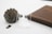 A pair of skull-themed earrings displayed on a transparent stand is set against a backdrop of a large pinecone and a dark wooden surface. The scene appears minimalist with a clean, white background, emphasizing the earthy tones of the wood and the textured pinecone.