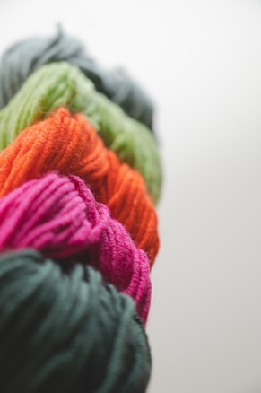 Bundles of yarn in vibrant pink, orange, green, and dark gray are arranged in a row. The texture of the yarn is soft and fibrous, with strands tightly wound together.