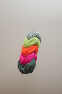 A neatly arranged bundle of colorful yarns is pictured against a plain background. The yarns are stacked vertically and consist of green, orange, pink, and dark green shades.