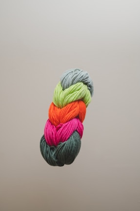 A neatly arranged bundle of colorful yarns is pictured against a plain background. The yarns are stacked vertically and consist of green, orange, pink, and dark green shades.