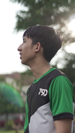 A young man with short dark hair is standing outdoors, wearing a sports shirt with green, black, and white colors. Sunlight filters through the trees in the background, creating a soft halo effect.