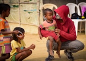 In a sunny outdoor setting, a seated person wearing a red hoodie and jeans is holding a small child in their lap. The child appears to be listening intently. Two other children, one with short hair in an orange striped shirt and the other with longer hair wearing a yellow shirt, are engaging nearby with playful expressions. The ground is sandy and several white plastic chairs are visible in the background, alongside a tent-like structure.