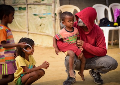 In a sunny outdoor setting, a seated person wearing a red hoodie and jeans is holding a small child in their lap. The child appears to be listening intently. Two other children, one with short hair in an orange striped shirt and the other with longer hair wearing a yellow shirt, are engaging nearby with playful expressions. The ground is sandy and several white plastic chairs are visible in the background, alongside a tent-like structure.