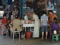 A small market or street vendor with several women interacting around a table displaying various goods, including packaged food items and bottled products. The vendor is situated in a busy area, with posters and shelves filled with merchandise in the background. The atmosphere is casual, with women wearing a variety of colorful clothing styles.