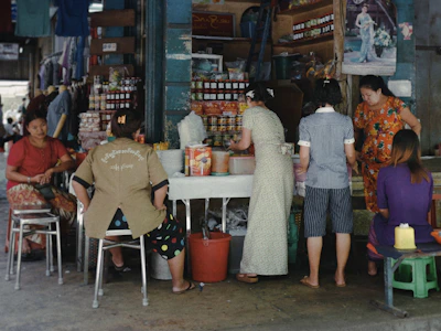 A small market or street vendor with several women interacting around a table displaying various goods, including packaged food items and bottled products. The vendor is situated in a busy area, with posters and shelves filled with merchandise in the background. The atmosphere is casual, with women wearing a variety of colorful clothing styles.