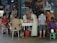 A small market or street vendor with several women interacting around a table displaying various goods, including packaged food items and bottled products. The vendor is situated in a busy area, with posters and shelves filled with merchandise in the background. The atmosphere is casual, with women wearing a variety of colorful clothing styles.