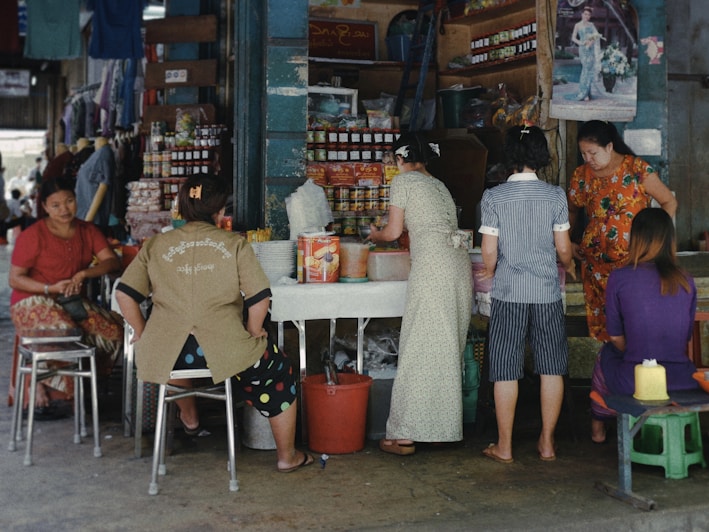 A small market or street vendor with several women interacting around a table displaying various goods, including packaged food items and bottled products. The vendor is situated in a busy area, with posters and shelves filled with merchandise in the background. The atmosphere is casual, with women wearing a variety of colorful clothing styles.