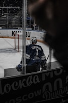A hockey player wearing a blue jersey with the name 'Yeryomko' and number 1 stands on the ice rink near a red goalpost. The player is seen from behind, and the surrounding area includes advertisements on the boards. The atmosphere appears to be inside an arena with a focus on the player.