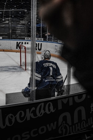 A hockey player wearing a blue jersey with the name 'Yeryomko' and number 1 stands on the ice rink near a red goalpost. The player is seen from behind, and the surrounding area includes advertisements on the boards. The atmosphere appears to be inside an arena with a focus on the player.