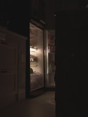 An open refrigerator illuminates a dimly lit kitchen. The light from the fridge reveals various food items and containers inside, casting soft shadows around. The surrounding area is predominantly dark, adding a contrast to the bright interior of the fridge.