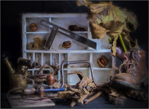 A rustic and nostalgic scene featuring old tools, dried leaves, and chestnuts set against a vintage-looking wooden shelf. The objects include a shoe, a saw, other woodworking tools, and a brass kettle. The setting suggests an autumnal theme with a mix of crafted and natural elements.