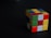 A partially solved Rubik's Cube with various colors including red, green, yellow, and blue on a dark surface. The cube's orientation emphasizes its three-dimensional shape, with a slight shadow cast.